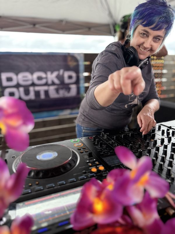 Jen Woolfe pointing at the camera from behind the decks, with pink orchids in the foreground